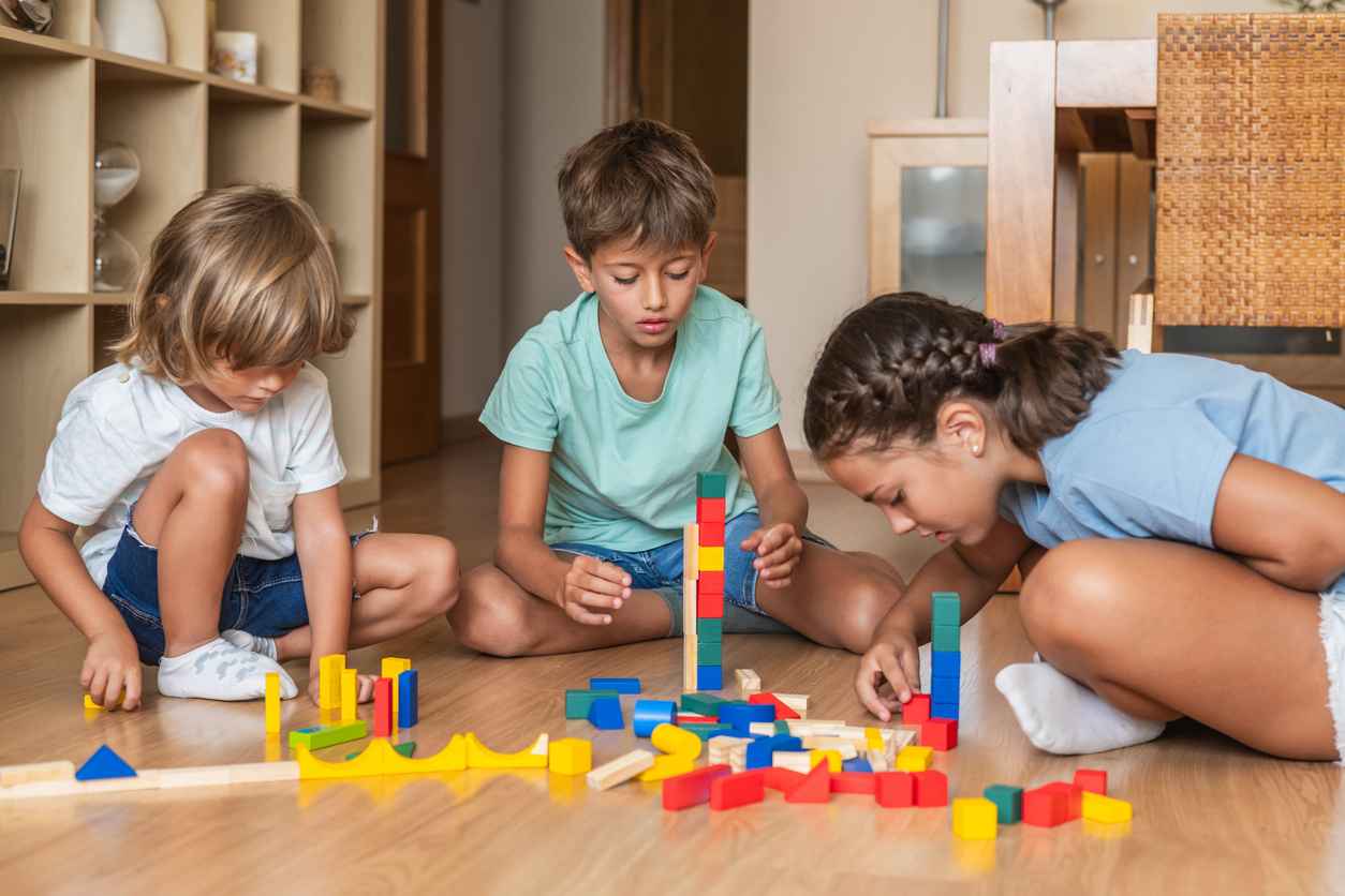 Family Time, Early Childhood Education, And Fun At Home. Three Happy And Focused Children Sitting On The Floor, Playing And Building With Wooden Blocks.